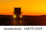 CLOSE UP LENS FLARE: Semi truck driving and hauling goods on empty highway across the Great Plains in golden morning. Freight delivery truck transporting cargo on interstate freeway at stunning sunset
