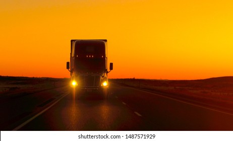 CLOSE UP LENS FLARE: Semi truck driving and hauling goods on empty highway across the Great Plains in golden morning. Freight delivery truck transporting cargo on interstate freeway at stunning sunset