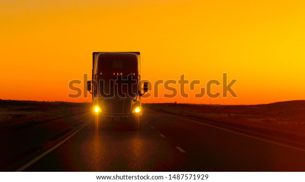 CLOSE UP LENS FLARE: Semi truck driving and\
hauling goods on empty highway across the Great Plains in golden\
morning. Freight delivery truck transporting cargo on interstate\
freeway at stunning\
sunset