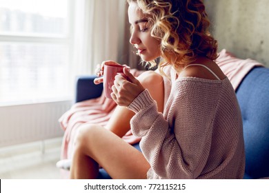 Close Up Lifestyle Portrait Of   Pensive Woman Drinking   Coffee In Early Morning, Sitting On Coach In Pink Cozy Sweater. 