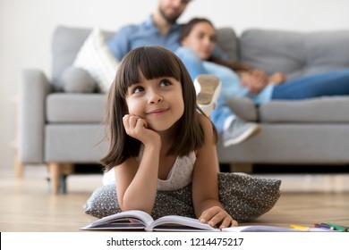 Close Up Little Adorable Thoughtful Smiling Daughter Dreaming Lying At Cushion On Warm Floor With Book In Living Room At Modern Home, Resting Married Couple Parents On Background, Focus On Small Kid