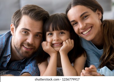 Close up little cute daughter holding hands on chin lying on warm floor with young parents in living room at home married couple sweet child smiling and looking at camera. Happy diverse family concept