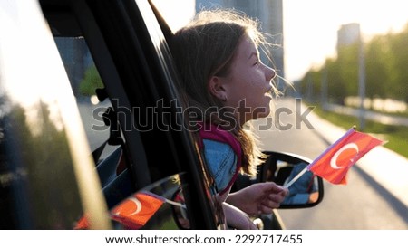 Close up of little cute girl in car window holding Turkish flag in hands. Beautiful child looking out of vehicle window in sunshine going on family trip. Small kid with flag in car. Patriotic concept