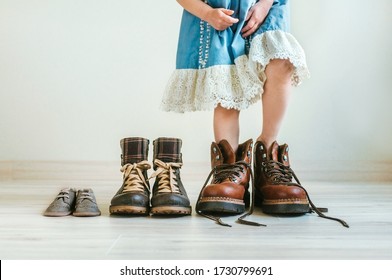 Close Up Of Little Girl In  Dress Putting On Father's Hiking Shoes. Father's, Mother's And Kids Boots Standing In A Row Indoor. Family Concept. Selective Focus On The Boots.