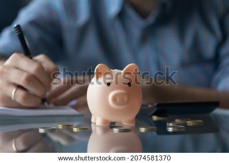 Close up little piggybank with young man writing notes in copybook on background, monthly budget managing, planning investment, savings for medical insurance or study, financial literacy concept.