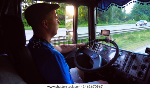 Close\
up of lorry driver riding through countryside. Man in hat\
controlling his truck and carefully watching traffic. Bright sunset\
light reflecting in side mirror of car. Slow\
motion