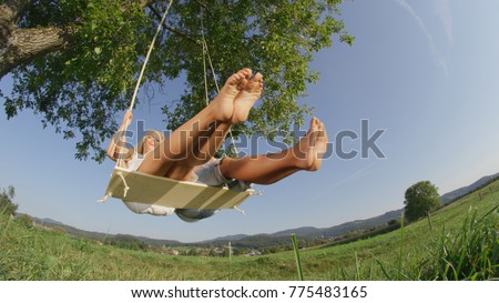 CLOSE UP LOW ANGLE: Barefoot couple with outstretched legs swinging over camera in sunny green pasture. Unrecognizable young couple on date swaying on wooden rope swing under large old tree in nature.
