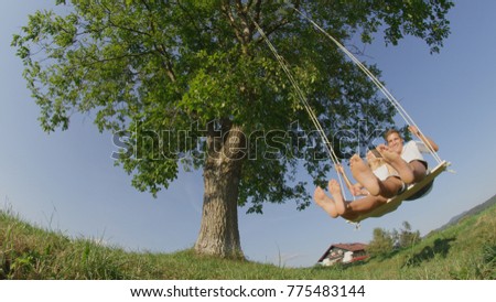 CLOSE UP LOW ANGLE Beautiful couple swinging under big tree in countryside in summer. Smiling girlfriend and boyfriend on a romantic date swaying barefoot in romantic rural setting on sunny afternoon.