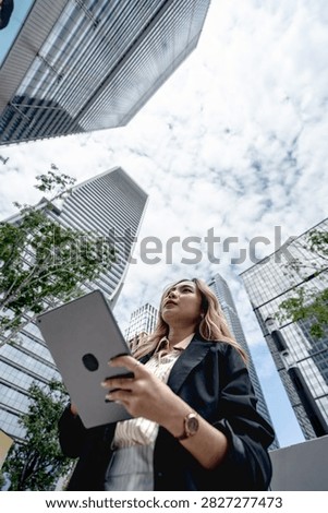 Close low angle view of an Asian professional in a black blazer holding her tablet near her chest while tilting her head far back to study the towering glass buildings above her.