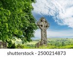 Close up low angle view of celtic cross in graveyard of the St Pancras Church Widecombe in the Moor, UK with leaves of green tree in front in Dartmoor National Park against white clouded blue sky