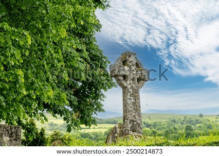 Close up low angle view of celtic cross in graveyard of the St Pancras Church Widecombe in the Moor, UK with leaves of green tree in front in Dartmoor National Park against white clouded blue sky