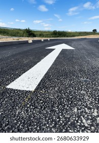 Close up low angle view of a large white directional arrow painted on a fresh asphalt road under blue sky with green grass on roadside and bright daylight