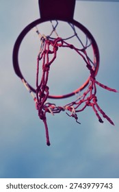 Close up low angle view looking directly through a red street basketball hoop with a torn and tangled red net against a soft cloudy sky