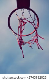 Close up low angle view of a weathered red street basketball hoop with a torn red net against a cloudy blue sky