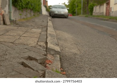 Close low-angle view of a deteriorated concrete curb and uneven paved sidewalk next to an asphalt residential road. Foreground shows broken paving blocks