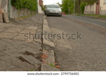 Close low-angle view of a deteriorated concrete curb and uneven paved sidewalk next to an asphalt residential road. Foreground shows broken paving blocks