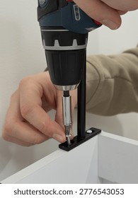 Close up macro view of cordless drill with chrome magnetic bit holder driving screw into small black metal bracket on top edge of white shoe cabinet panel