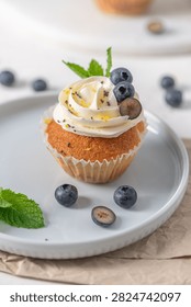 Close up macro view of a fluffy cupcake featuring swirled white frosting, poppy seeds, and bright natural lighting, garnished with sliced blueberries, fresh mint, and lemon zest.