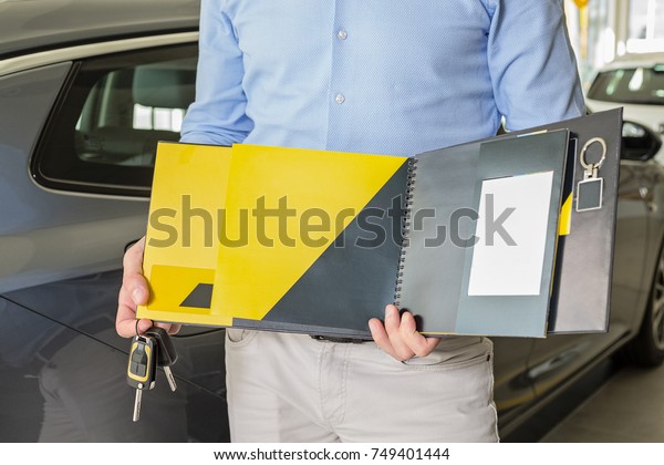 Close up of male adult\
car dealer standing indoors at car dealership, holding an opened\
blank yellow and black booklet and car keys in his hands, cars at\
the background