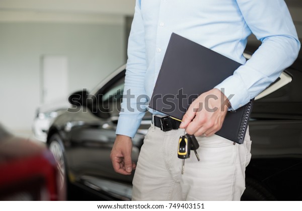 Close up of male adult car dealer standing indoors at\
car dealership, holding a folder and car keys in his hand with a\
new car behind him