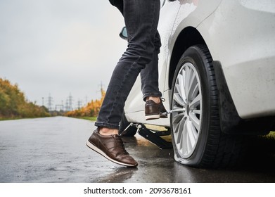 Close up of male auto mechanic standing on metal wrench while unscrewing lug nuts on car wheel. Young man using lug nut wrench while changing flat tire on the road. Concept of emergency road service.