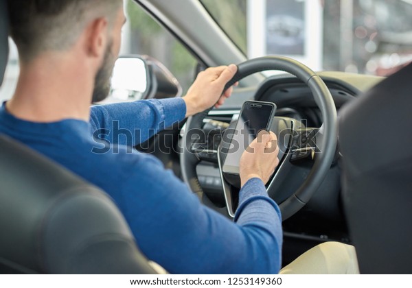 Close up of male back sitting in front seat of car
and using smartphone. Brunette man testing new luxury auto in
salon. Young driver holding one his hand on steering wheel and in
other having phone.