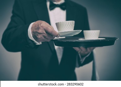 Close Up Of Male Butler Wearing Formal Suit And Bow Tie Carrying Tray Of White Coffee Mugs And Serving Cup Towards Camera