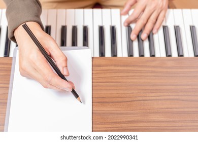 close up male composer hand writing a song on white blank music sheet while playing acoustic piano
