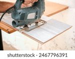 Close up of male construction worker hands cutting laminate wooden board with jigsaw tool. Man using electric power saw cutter while preparing laminate material for floor installation