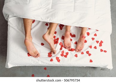 Close Up Of Male And Female Feet. Loving Couple Is Lying On Bed Under Blanket Covered By Small Red Paper Hearts