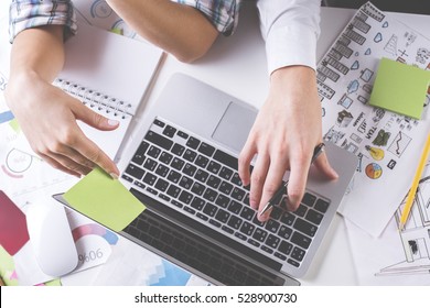 Close Up Of Male And Female Hands Using Laptop Placed On Messy Desktop With Financial Reports And Supplies