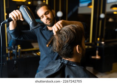 Close up of a male hairstylist using a blow dryer and a brush after cutting the hair of a man customer at the salon