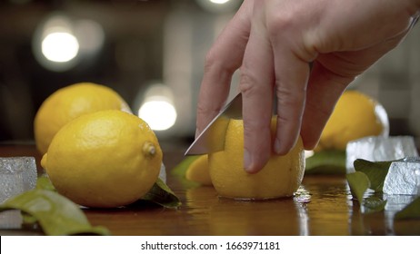 Close up male hand cutting lemon into wedges on the table over background of blurry light bulbs. Cooking of refreshing drinks. Bartender working closeup