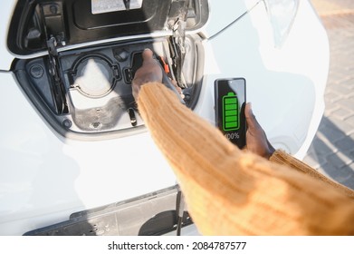 Close up of male hand holding electric charging plug for luxury red car. African businessman charging his auto on station. Eco friendly transport. - Shutterstock ID 2084787577