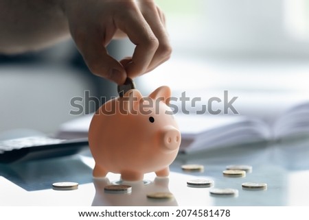 Close up male hand putting coins inside small piggybank, young man collecting cash saving money for future purchases, planning investments, managing household budget expenditures, financial literacy.