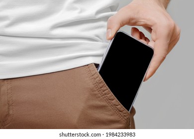 Close up of male hand taking out big mobile phone with empty black screen from front pocket of brown pants on grey background.