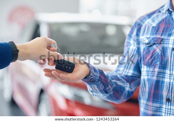 Close up of male hands and body in blue\
checkered shirt. Crop of hands when one person giving car\'s key to\
another. Man taking key from auto from his friend. On background\
standing red vehicle.