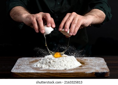 Close Up Of Male Hands Cracking Eggs Upon Flour, Making Dough For Bread Or Pastry. Baker Man Wearing A Black Outfit In Darkness.