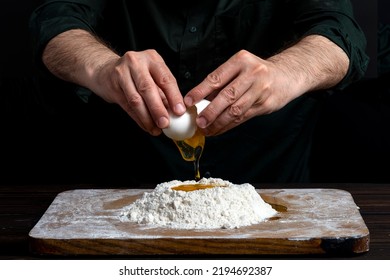 Close Up Of Male Hands Cracking Eggs Upon Flour, Making Dough For Bread Or Pastry. Baker Man Wearing A Black Outfit In Darkness.