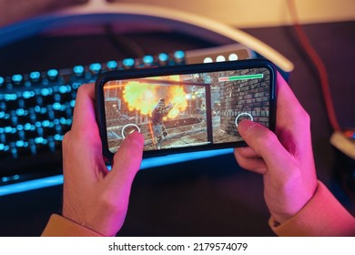 Close up of male pro gamer's hands holding smartphone with online video shooter game. Cyber sportsman playing mobile games on his cellphone. Professional keyboard on background. Top view. Neon lights
