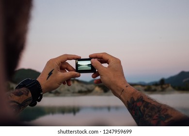 Close up of male tattooed hands and arms hold action camera and make photo f sunset or sunrise during amazing adventure or travel road trip. Inspirational outdoors wanderlust motivation