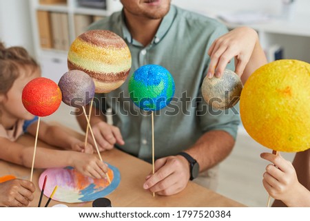 Close up of male teacher holding planet models and looking at camera while working with group of children in art and craft lesson, copy space