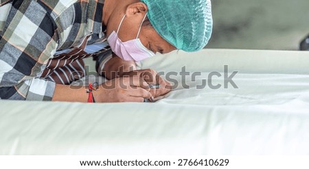 Close up of male textile worker using magnifying tool to check fabric quality inspection in factory. Asian supervisor in mask with magnifier inspecting thread details for production accuracy.
