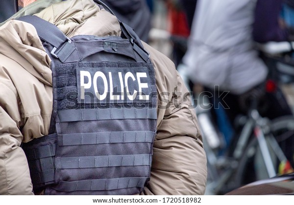 A close up of a male undercover police officer stands on\
a street among a crowd of people. The officer has a black canvas\
bullet proof vest on with the word police spelled out in white\
letters. 