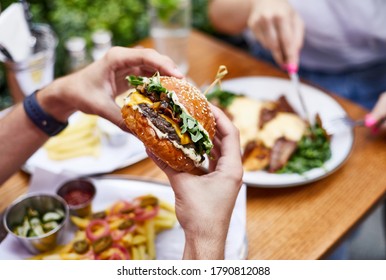 Close up male's hands holding delicious fresh burger. Young man eating tasty hamburger sitting at table in outdoor cafe on sunny summer day. - Powered by Shutterstock