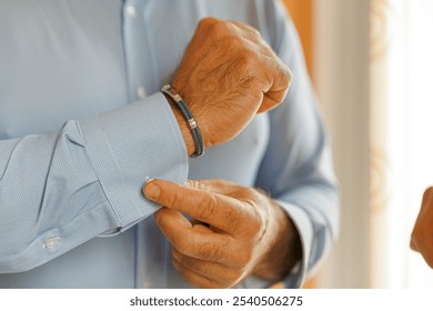 Close up of a man arm and hands while he tiles the cuffs of his shirt. Blue shirt with a green bracelet 