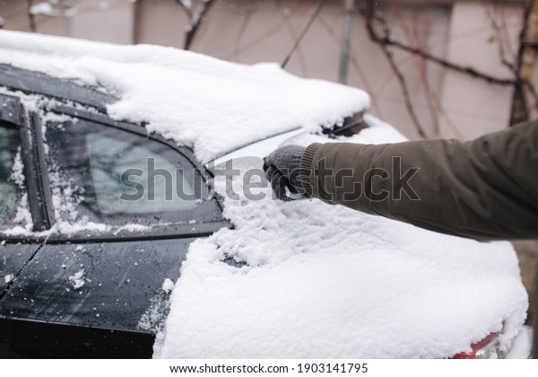 Close up of man is cleaning snowy window on a car\
with snow scraper. Focus on the scraper. Cold snowy and frosty\
morning. Black car