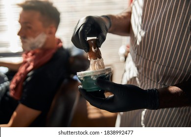 Close up of man hairdresser hands in sterile gloves dipping shave brush into bowl with cream while gentleman sitting in barber chair - Powered by Shutterstock