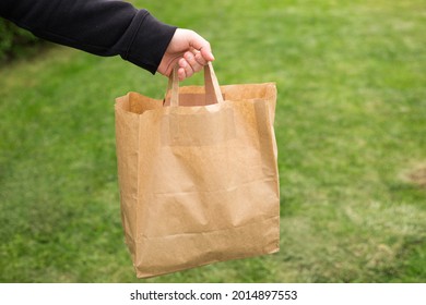 Close Up Of Man Hand With Eco Paper Bag For Takeaway Food On Nature Green Background. Delivery In Any Weather Around The Clock To The Client.