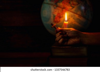 Close Up Of A Man Hand Holding Small Wooden Cross On Holy Bible Over Candle Light And Blurred World Globe In The Dark Room, World Mission Concept, Christian Background With Copy Space.